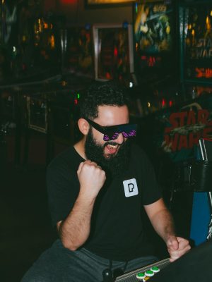 Cheerful man with sunglasses enjoying a classic arcade game in a vibrant arcade room.